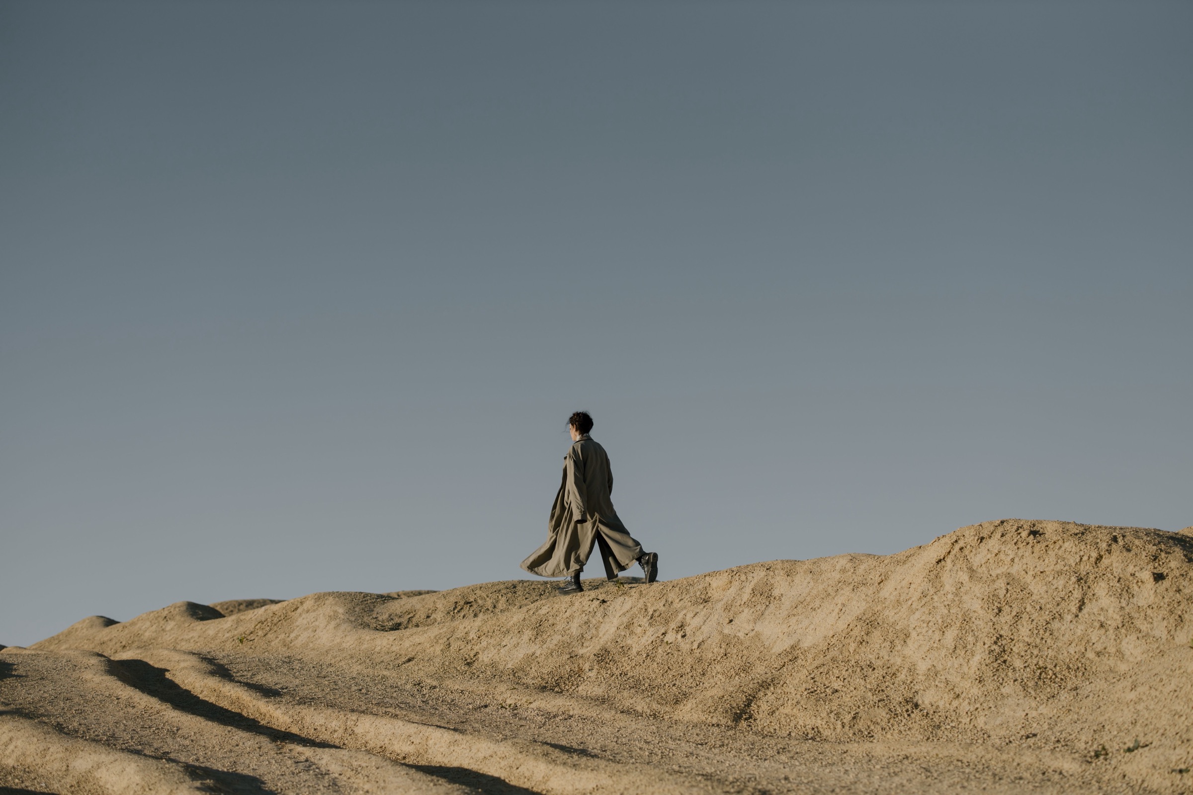 A model in an olive trench coat walks along the ridge of a desert dune at dusk